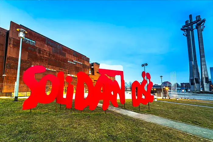 Den ikoniska röda Solidarność-logotypen utanför Solidaritetsmuseet i Gdańsk, med museets karakteristiska rostfärgade arkitektur och de tre korsen från monumentet över fallna varvsarbetare i bakgrunden.