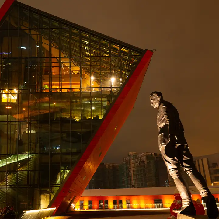 Den moderna arkitekturen hos Museet för andra världskriget i Gdansk upplyst nattetid, med en staty i förgrunden som blickar mot den lutande glasbyggnaden.