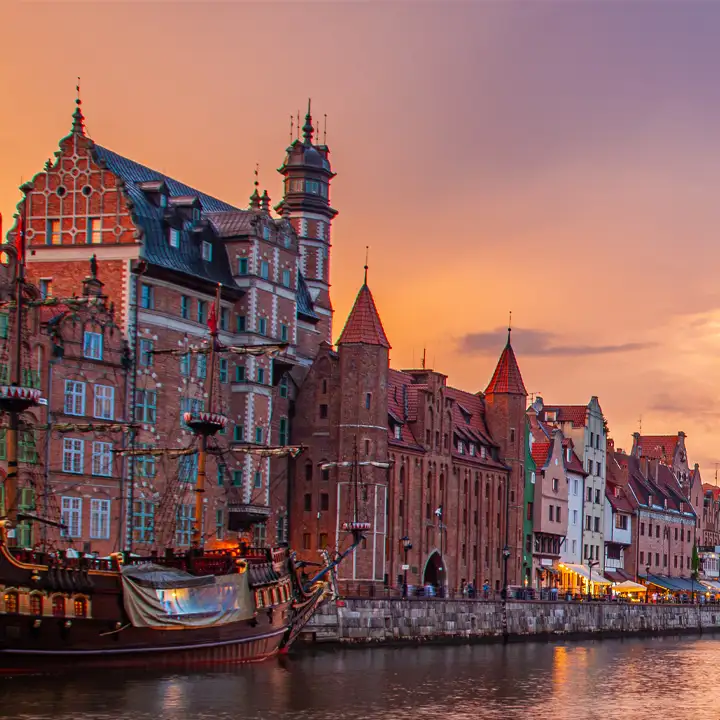 Ett gammalt piratskepp ligger förtöjt vid kajen längs floden Motława, framför de historiska husen vid Długie Pobrzeże i Gdansk under en vacker solnedgång.