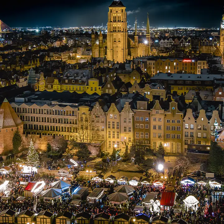 En stämningsfull vy från ovan över den upplysta julmarknaden i Gdansk med sina små marknadsbodar, julgranar och Mariakyrkan som tornar upp sig i bakgrunden mot natthimlen.
